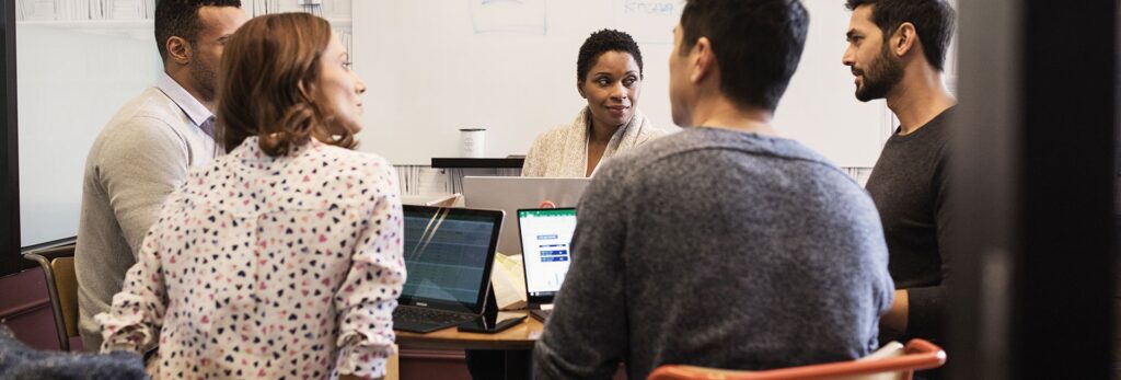 Five people sit around a table with laptops, having a discussion in a modern office with a whiteboard in the background.