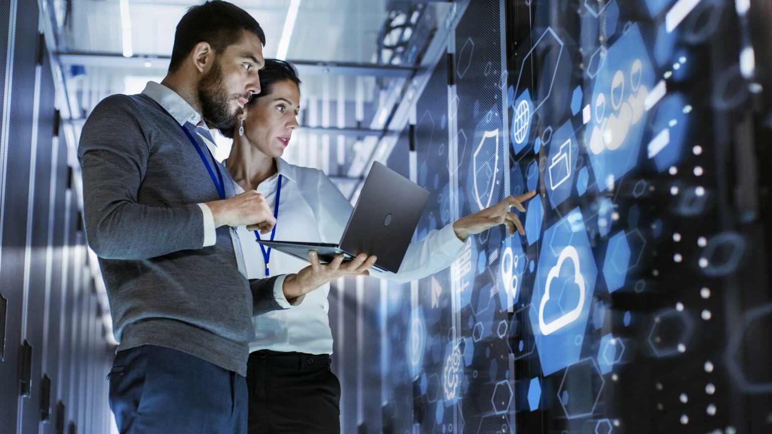 Two IT professionals discuss data displayed on a large digital screen in a server room, with one holding a laptop and the other pointing.