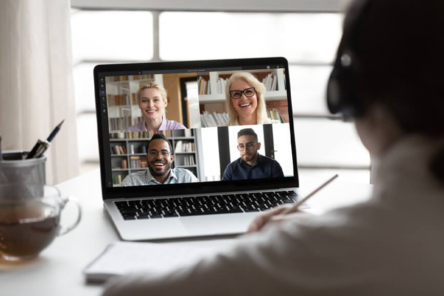 Person wearing headphones joins a video call with four people on a laptop, taking notes with a pencil at a desk.