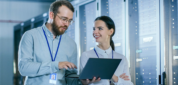 Two IT professionals with ID badges discuss something on a laptop in a server room filled with data server racks.