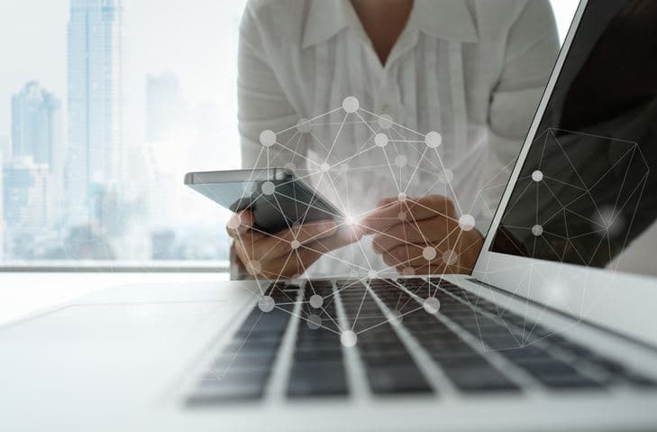 Person using a mobile phone and laptop with abstract digital network graphics overlaid, in a modern office setting.