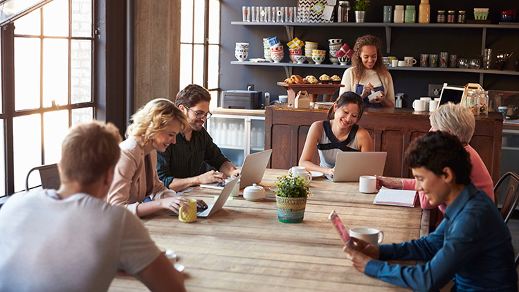 A group of people work on laptops and chat around a wooden table in a cosy café, with a barista preparing drinks in the background.