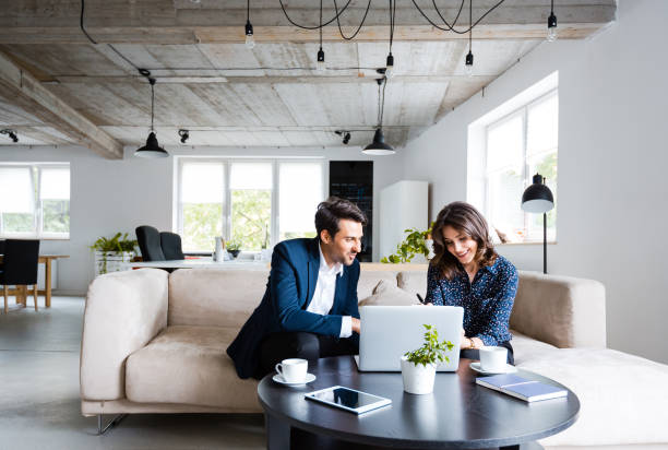 Two people sit on a sofa in a modern office, smiling and looking at a laptop on the coffee table in front of them.