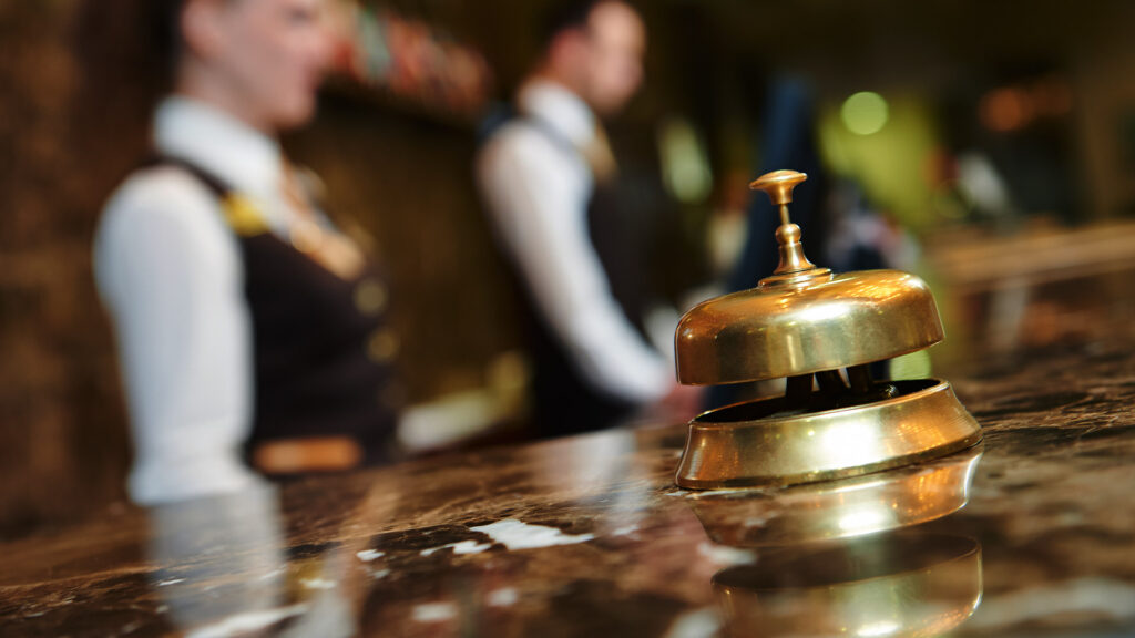 A hotel reception bell on a marble counter with two blurred staff members standing in the background.