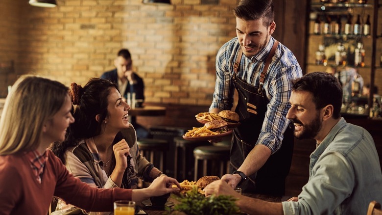 A waiter serves burgers and chips to three smiling friends sitting together at a restaurant table.