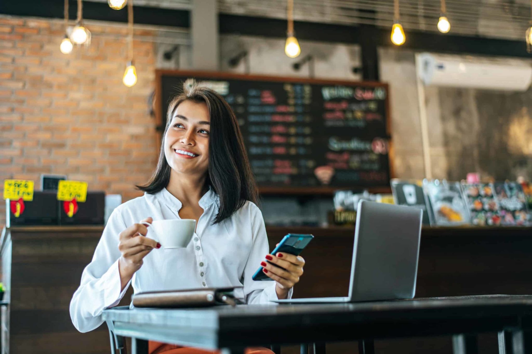 Smiling woman holding a cup and a mobile sits at a table with a laptop in a cosy café.
