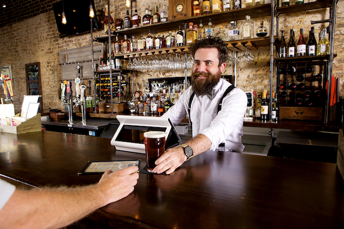 A smiling barman with a beard serves a pint of beer to a customer at a rustic bar with shelves of bottles behind him.