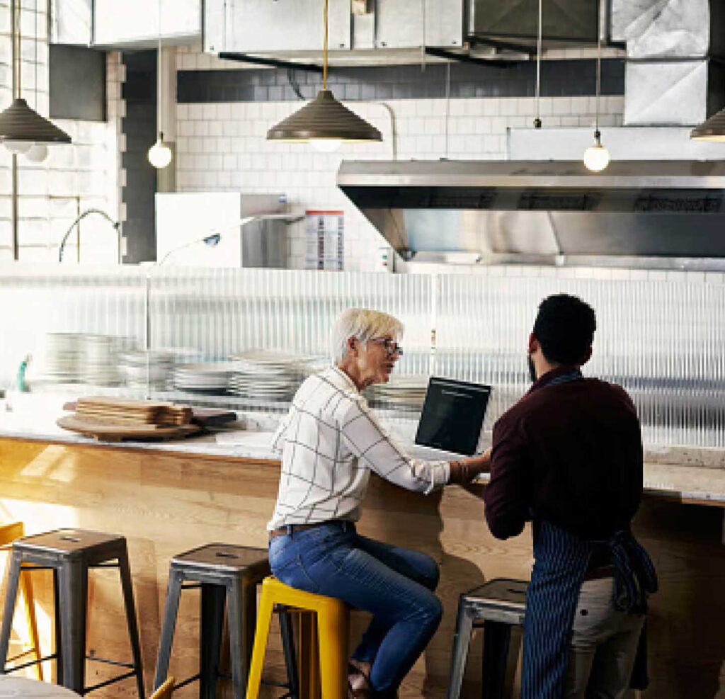 Two people sit at a counter in a modern restaurant kitchen, one using a laptop as they have a discussion.
