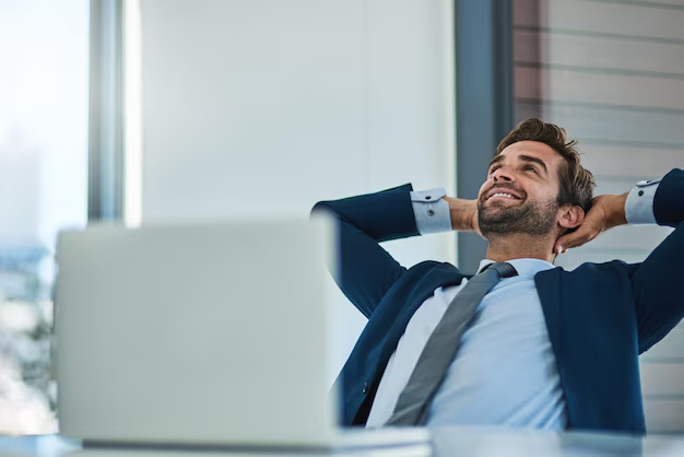 A smiling man in a suit leans back in his office chair with hands behind his head, looking relaxed, in front of a laptop computer.