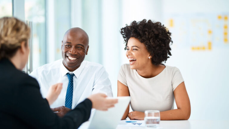 Three people sit at a table in a bright office, smiling and laughing whilst having a conversation.