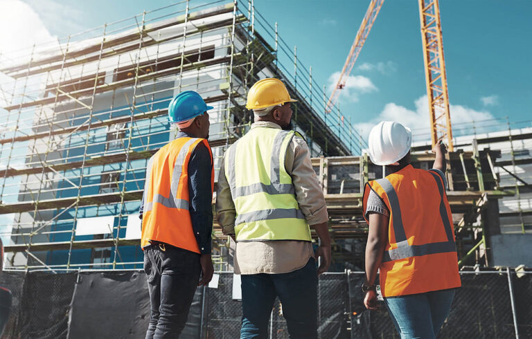 Three construction workers in safety gear look at a building under construction with scaffolding and a crane in the background.