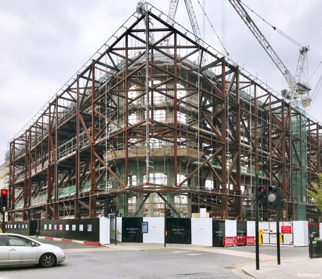 A large building under construction, with exposed steel framework and cranes above, is seen at a city crossroads with cars and traffic lights.