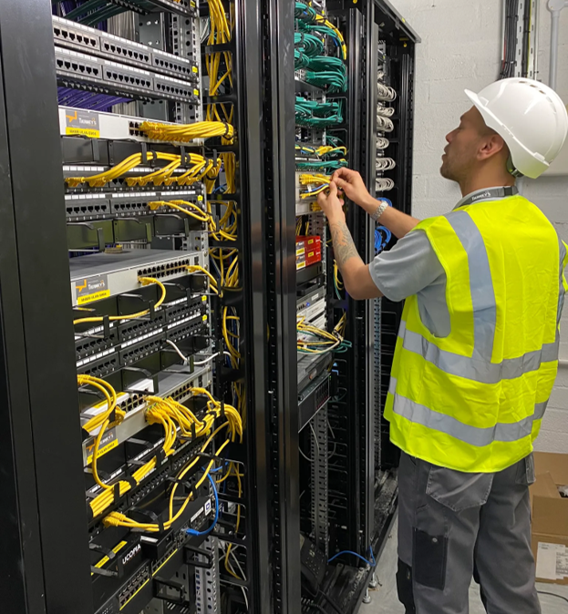 A worker in a hard hat and hi-vis vest manages cables in a server room filled with network racks.